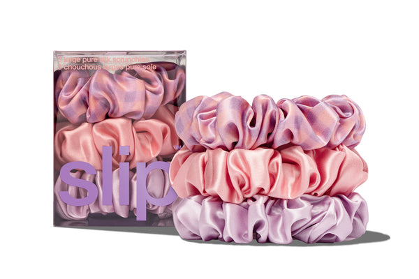 A set of three large scrunchies in shades of purple, pink, and rose, displayed alongside their packaging with the brand name 'slip' visible.