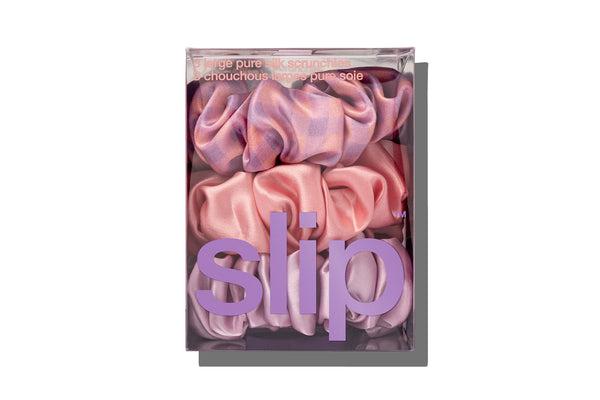 A set of three large slip silk scrunchies in shades of purple, pink, and rose in branded packaging on white background.