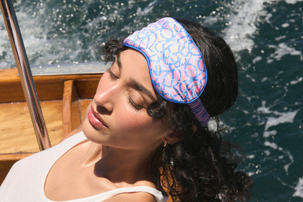 Woman on a boat wearing a slip silk sleep mask with a nautical rope themed pattern in blue and pink.