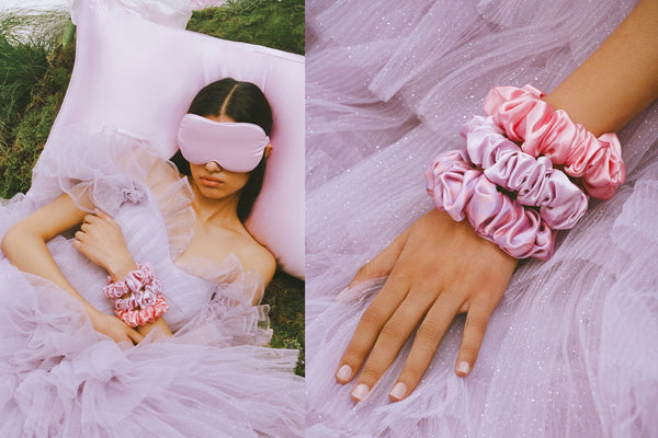 Woman lying down on a lilac slip silk queen pillowcase in a lilac dress, wearing a slip lilac contour mask and large scrunchies. Hand with three large pink and lilac slip scrunchies.