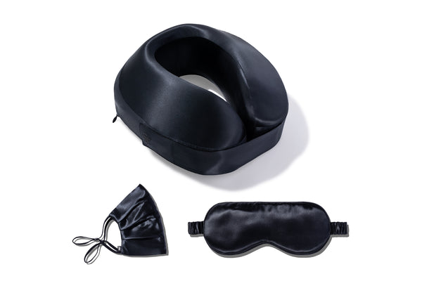 A travel pillow with a black pure silk removable pillowcase, a black pure silk sleep mask, and a black pure silk reusable face covering all laid out on a white background.