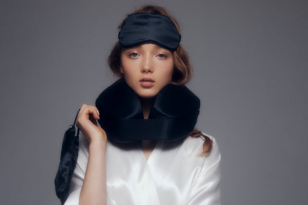 Model wearing a black silk travel pillow, a black pure silk sleep mask, and holding a black pure silk reusable face covering.