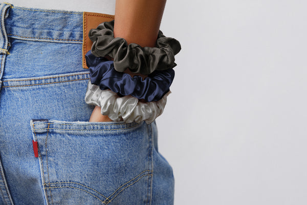 A model with hand in the back of denim shorts pocket. On the arm is three large silk scrunchies in charcoal, navy and silver.